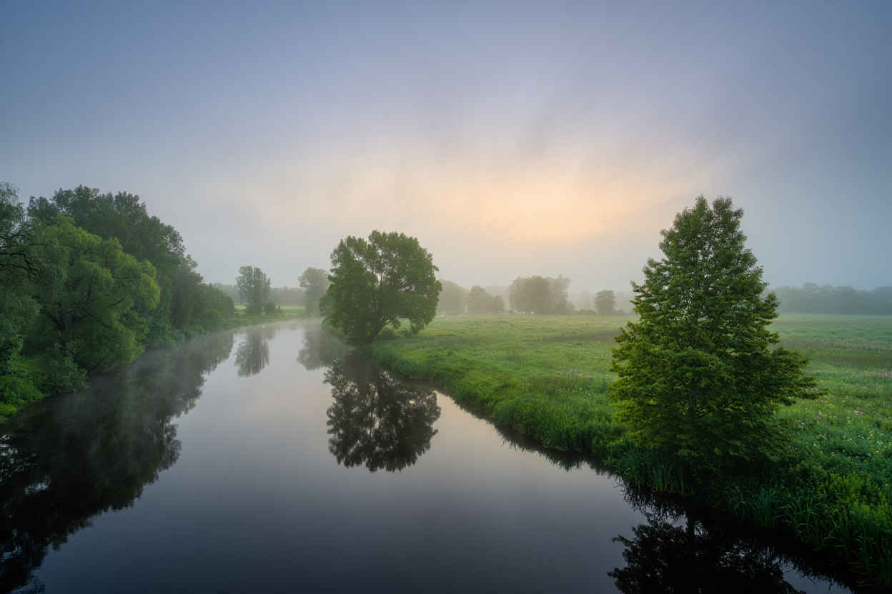 Stiller Fluss im Morgennebel umgeben von grünen Wiesen und sanften Hügeln in der deutschen Landschaft